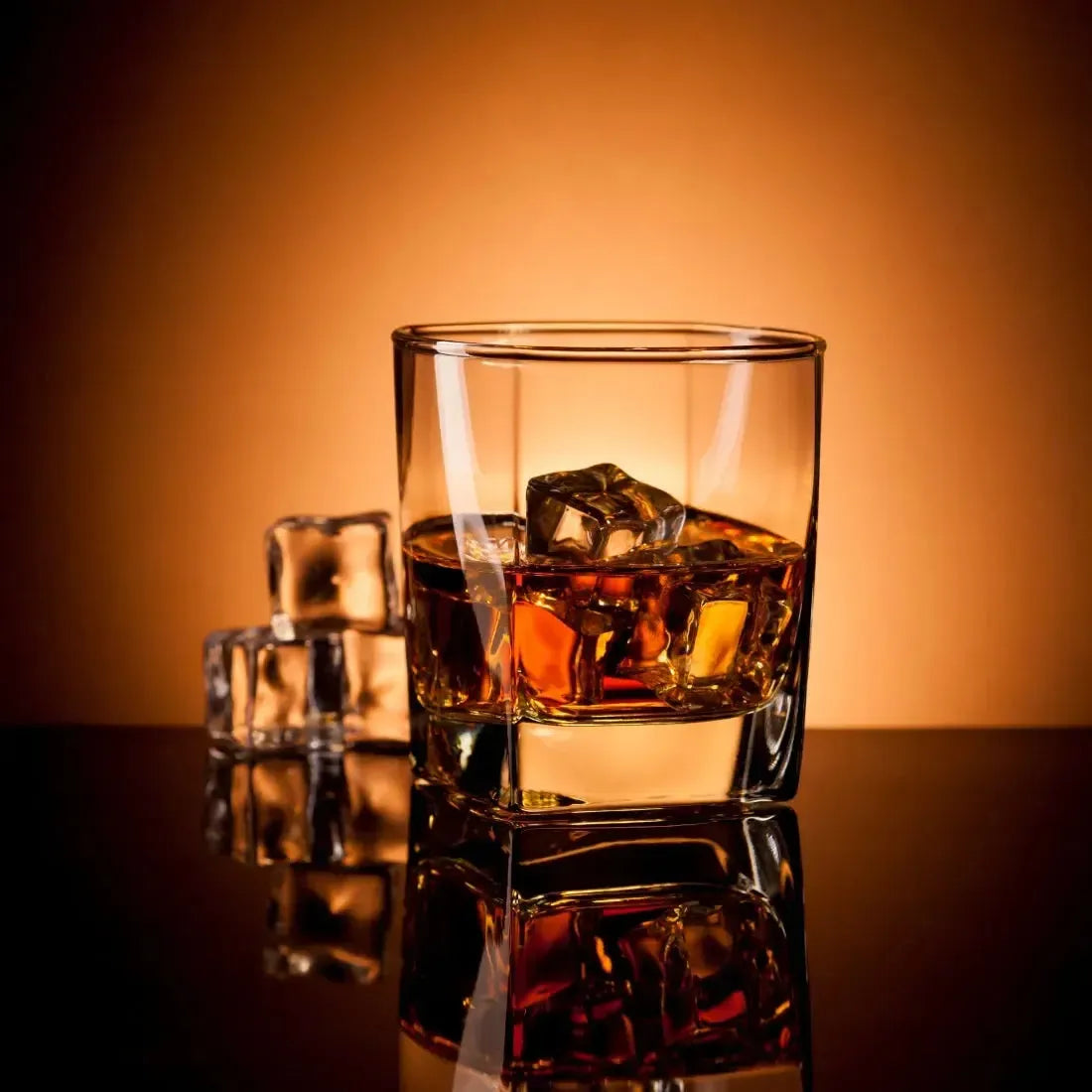 Glass of amber whiskey on the rocks with ice cubes against a warm orange backdrop.