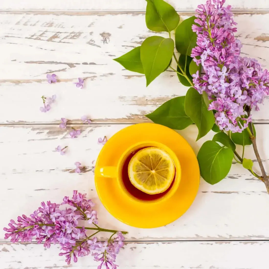 A cheerful yellow cup of tea with lemon, surrounded by blooming lilac flowers on a rustic wooden table.