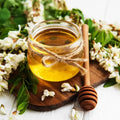 Jar of golden honey with a wooden honey dipper surrounded by fresh flowers and green leaves.