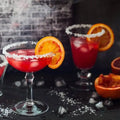 Vibrant orange cocktails with a salted rim and ice, garnished with fresh orange slices on a dark background.