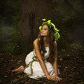 A serene woman dressed in white with a leaf crown, seated by a majestic tree in a mystical forest setting.