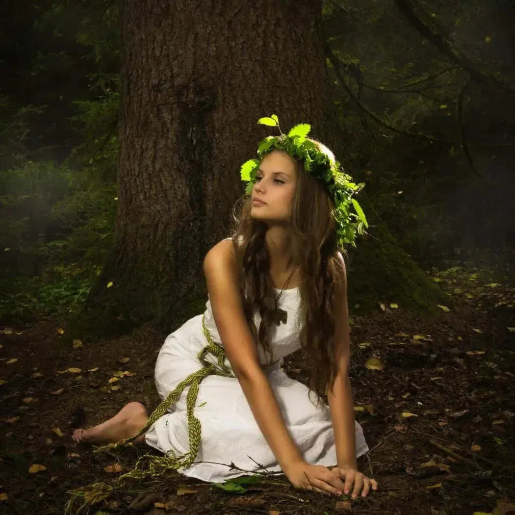A serene woman dressed in white with a leaf crown, seated by a majestic tree in a mystical forest setting.