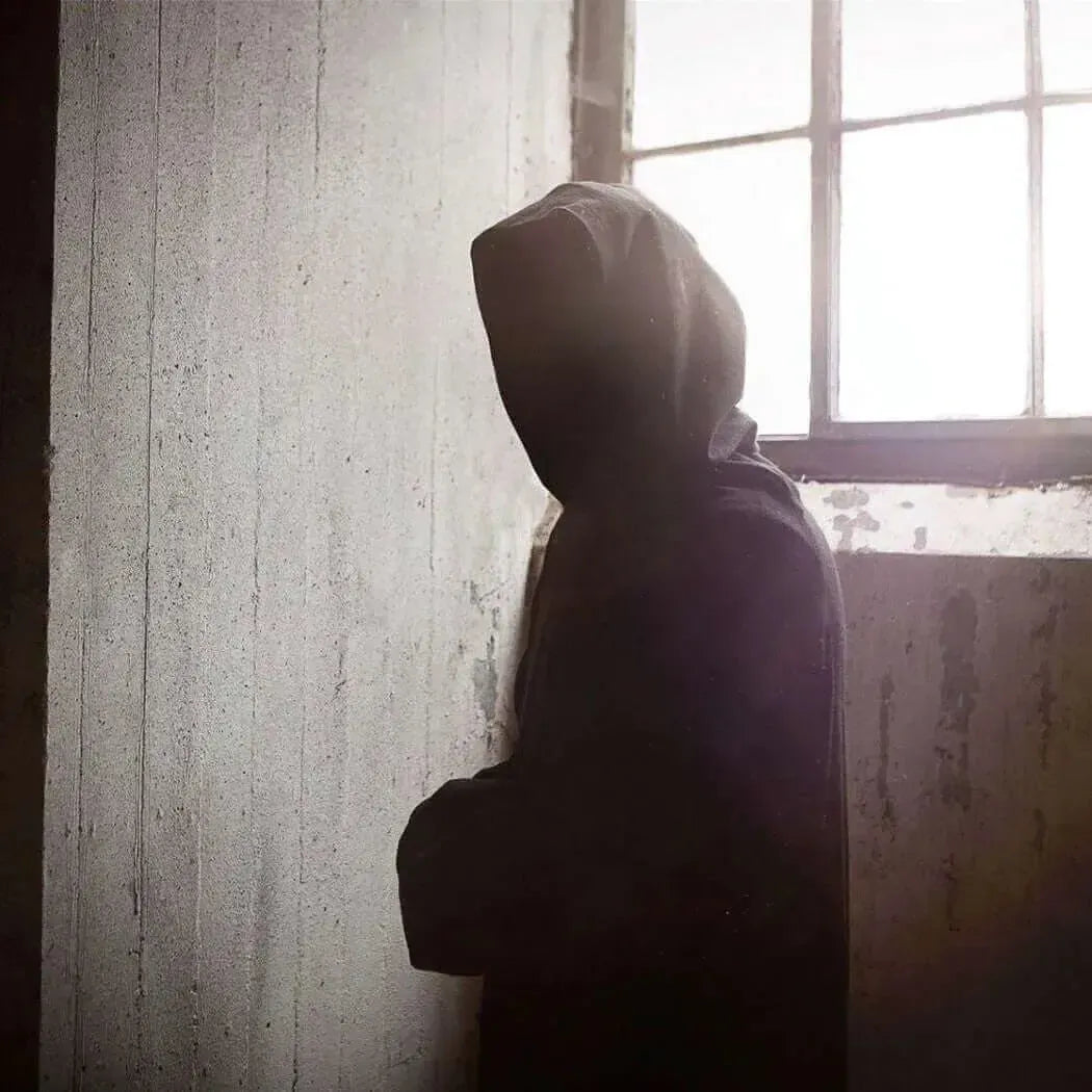 Mysterious figure wearing a hooded cloak, leaning against a wall near a window, embodying an enigmatic vibe.